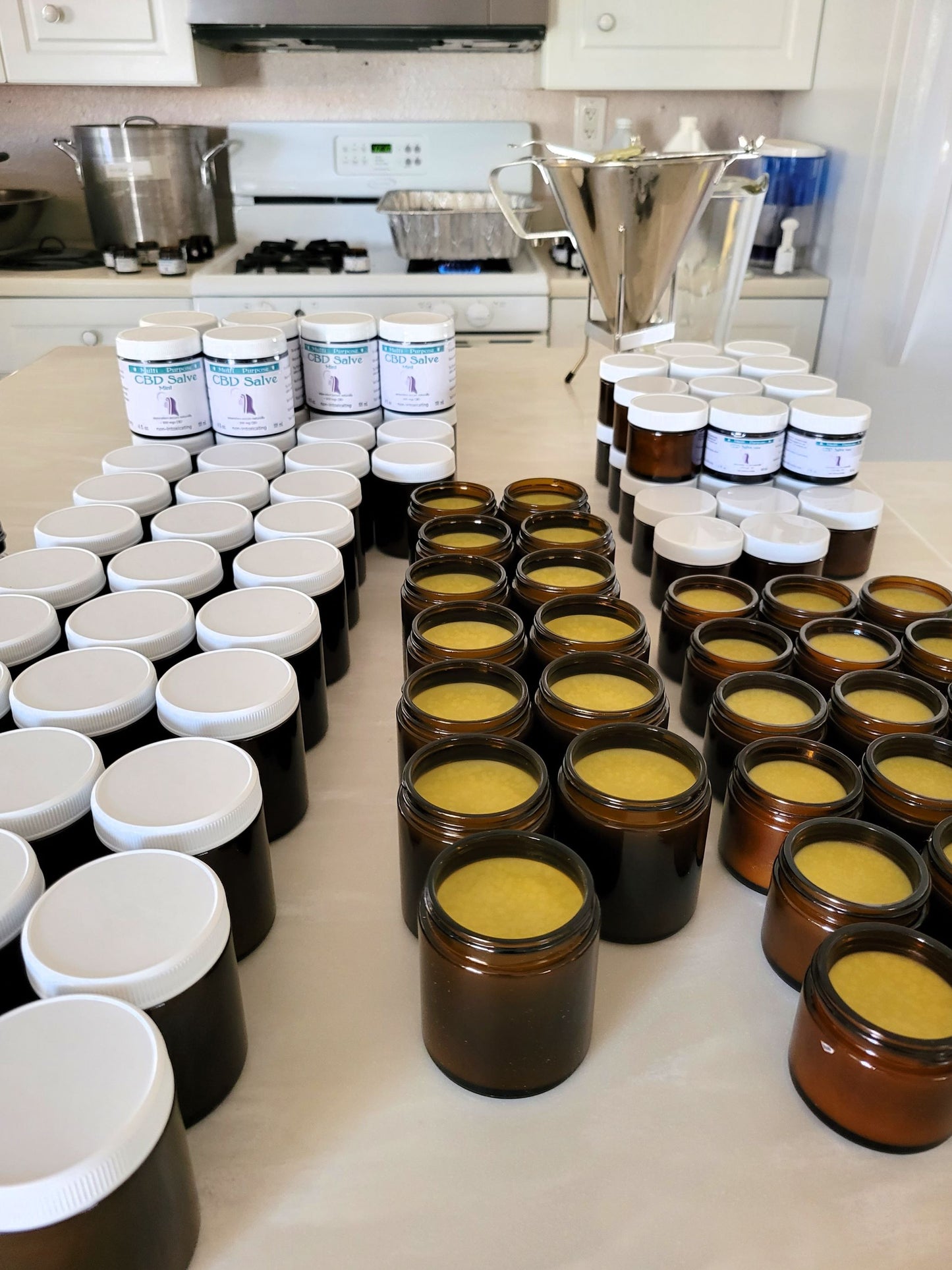 Rows of small glass jars filled with CBD Topical Salve 8 oz. Lavender or Mint (~1,000 mg CBD) sit on a kitchen counter; white-lidded jars, labeled containers, a stainless steel funnel, and appliances are also in view.